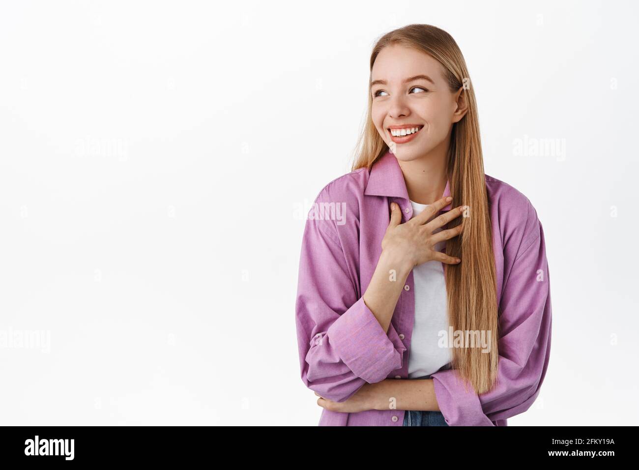 Smiling candid girl, looking aside, laughing with natural happy face emotion, touching chest, chuckle over something funny, standing against white Stock Photo