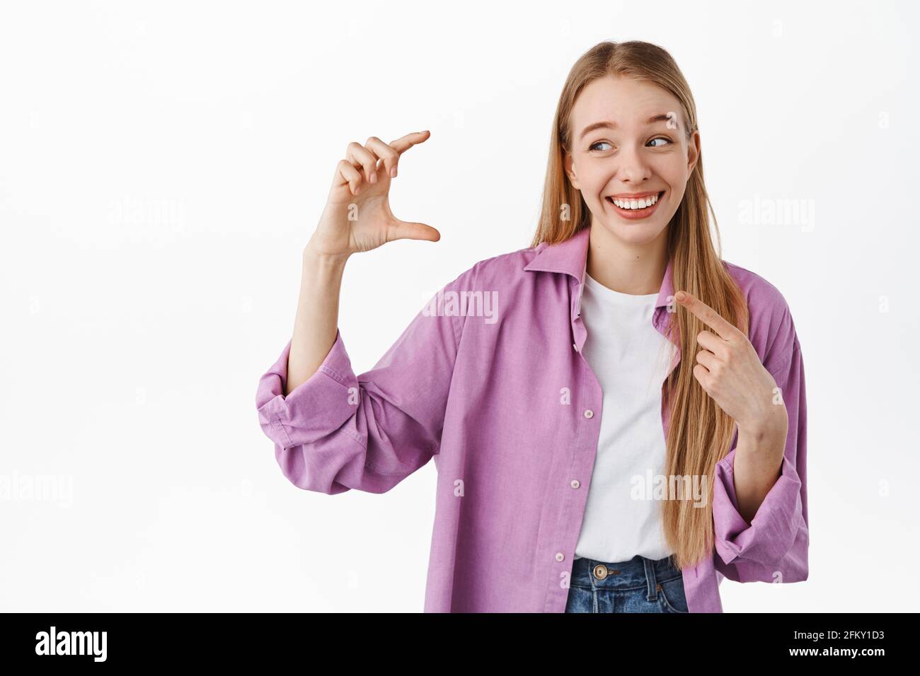 Blond woman holding something small and finy. Girl laughing while pointing at her hand showing little size tiny thing, smiling, standing against white Stock Photo