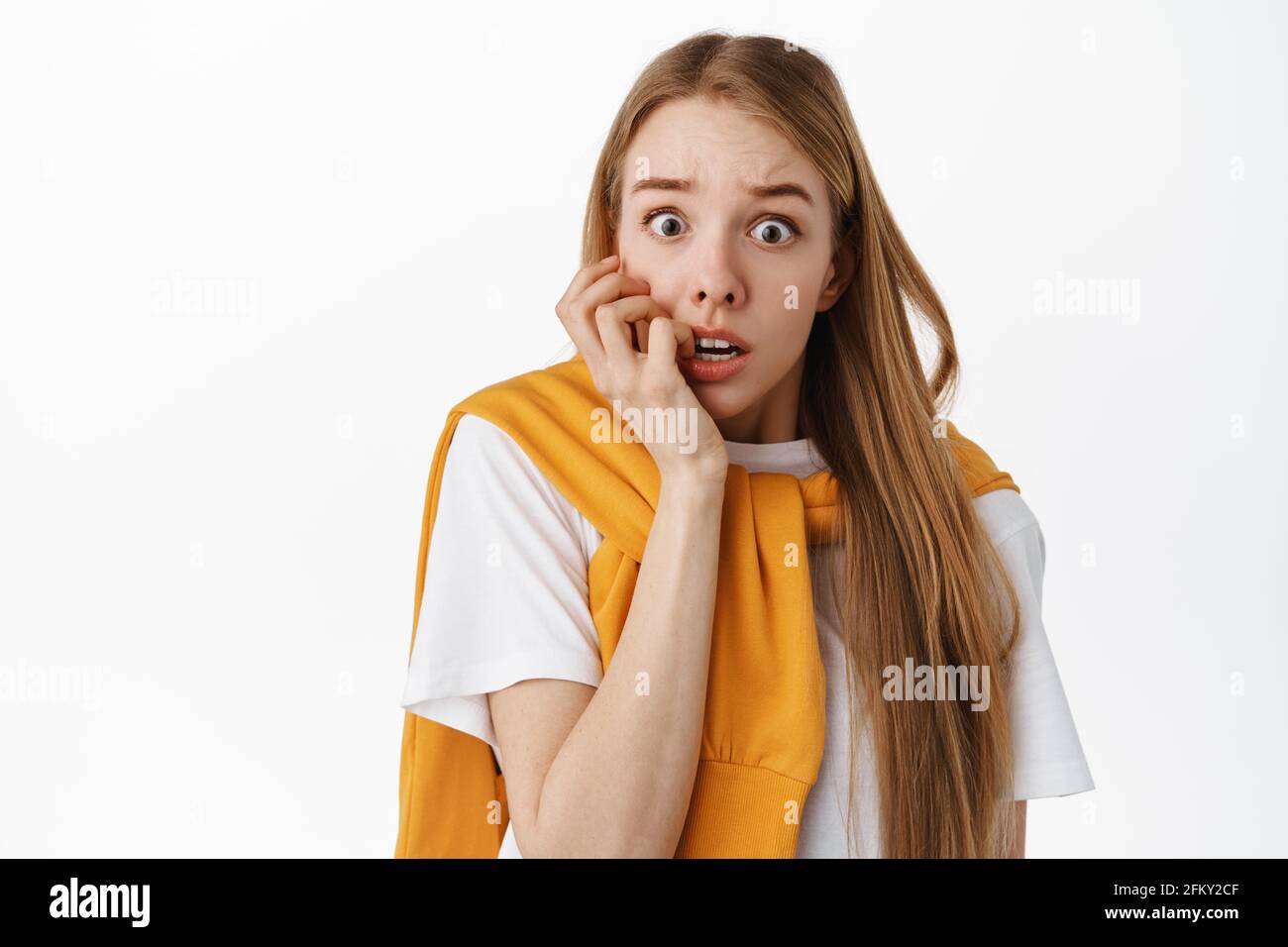 Close up of shocked and scared blond woman, biting fingers and gasping, staring startled and frightened, afraid of something scary, standing over Stock Photo