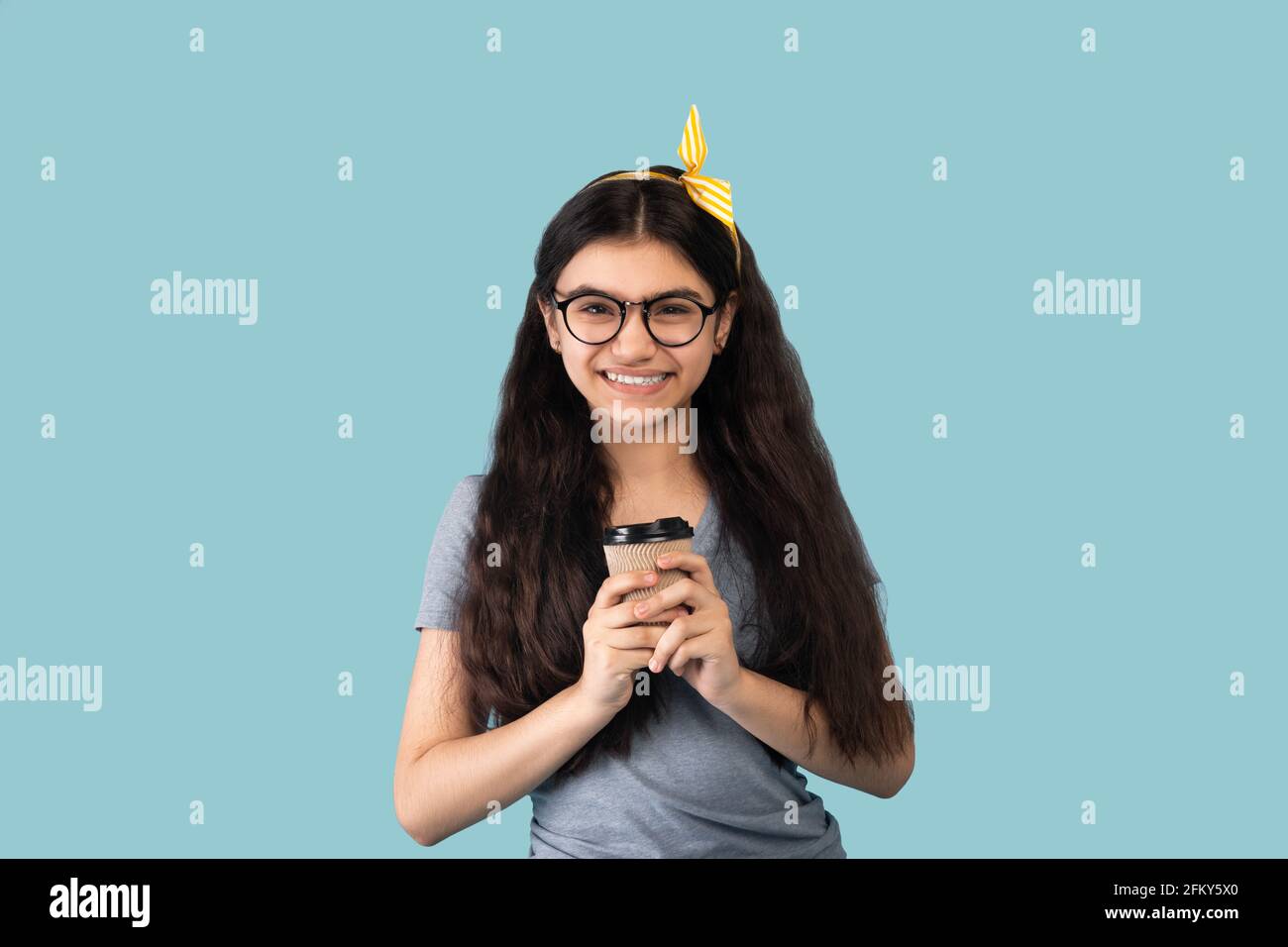 Happy Indian teen girl in casual clothes holding aromatic coffee in takeaway paper cup on blue background Stock Photo