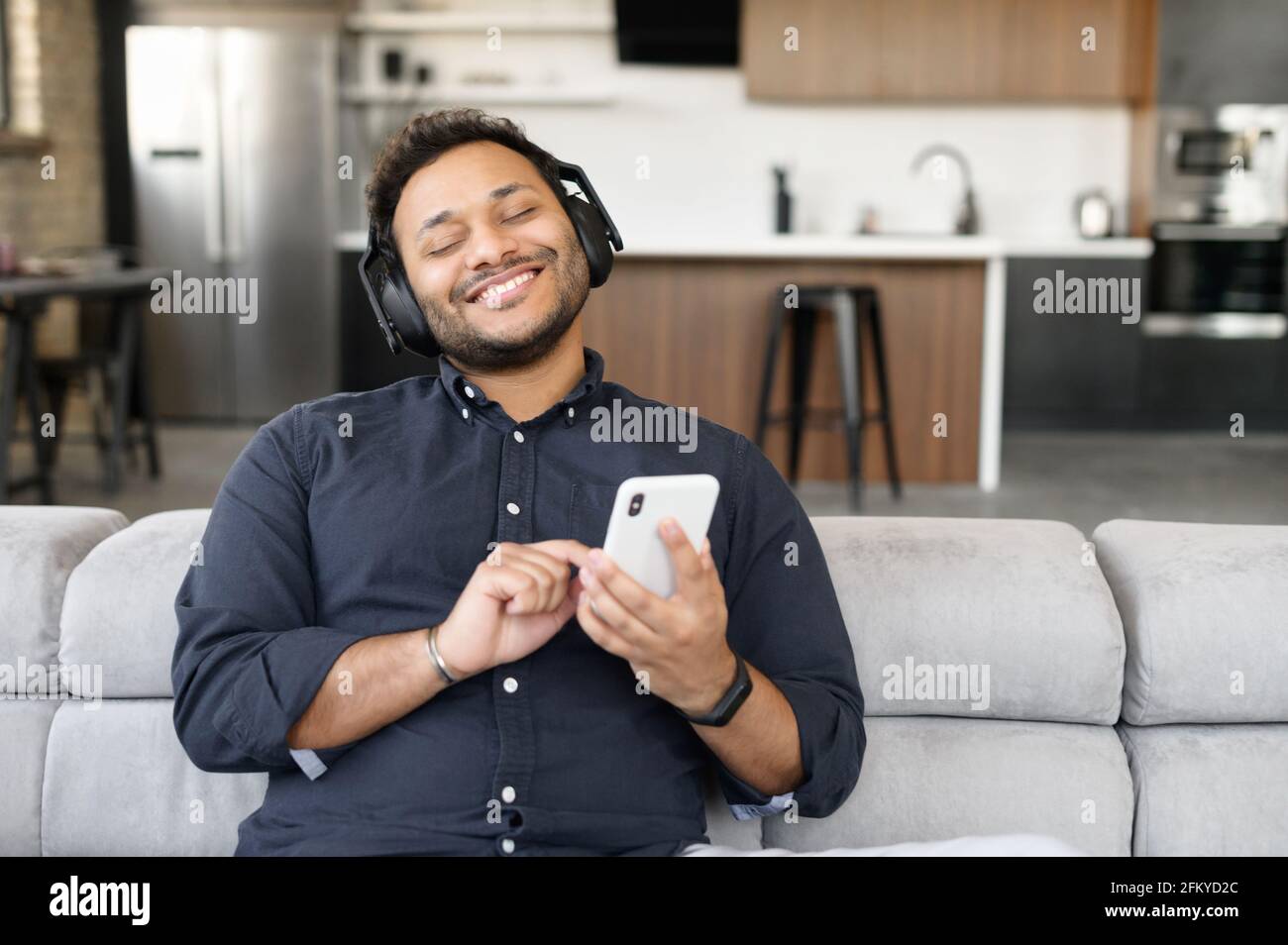 Cheerful handsome mixed-race guy wearing headset enjoys music sitting on the couch at home, indian man holding smartphone, listening favorite tracks with eyes closed, dreaming Stock Photo