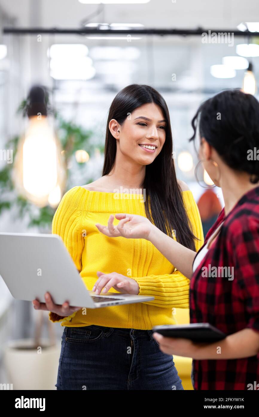 Beautiful girl with long dark hair working on laptop and discussing with her female colleague in a modern business office. Stock Photo