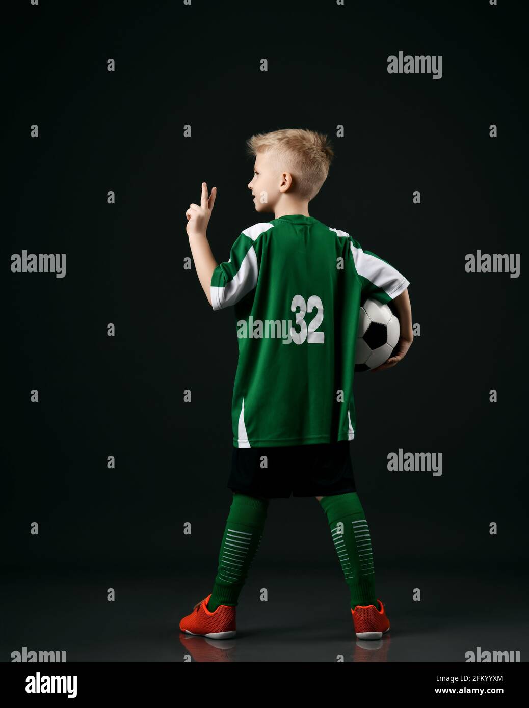 Soccer school student, teenager boy in red white striped uniform stands back to camera holding ball and gesturing V sign Stock Photo