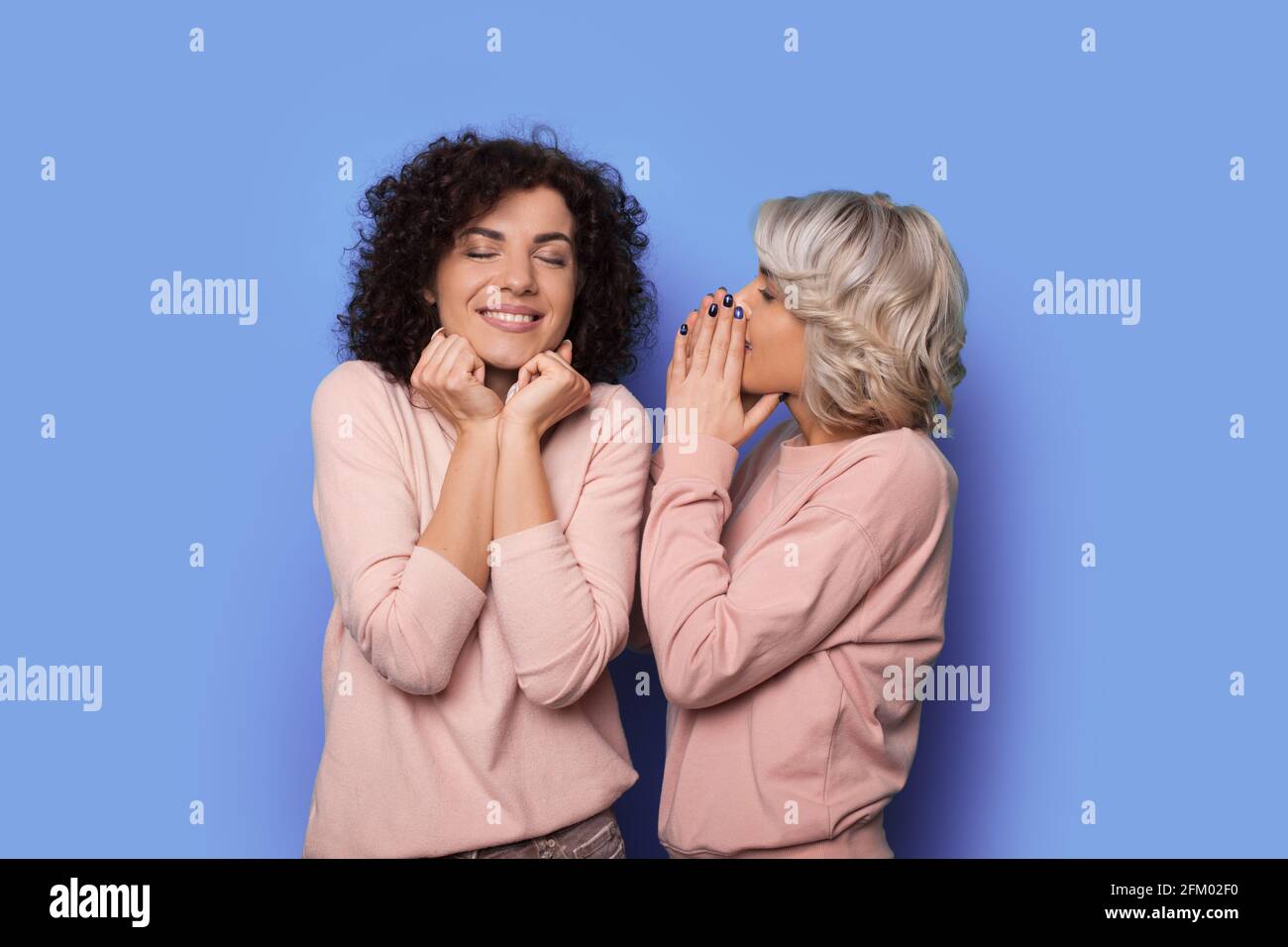 Blonde curly woman is whispering something to her brunette sister smiling on a blue studio wall Stock Photo