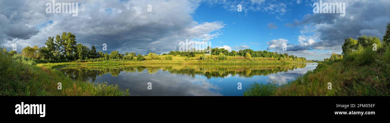 Stormy dramatic sky over the calm lake and green hills during sunset.Beautiful panoramic landscape. Stock Photo