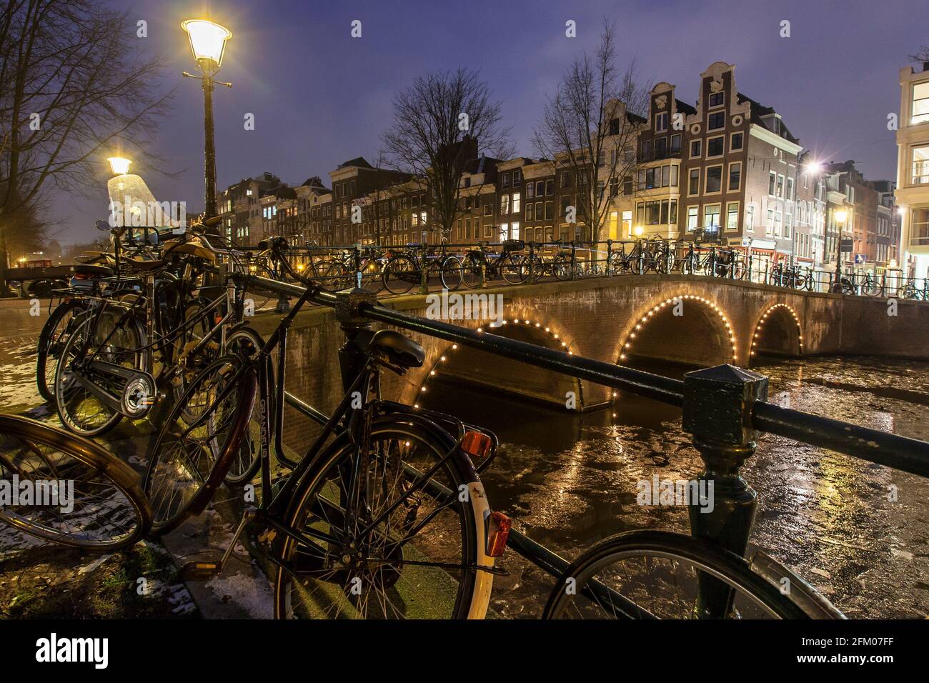 Bicycles parked on bridge on the icy Keizersgracht canal during winter dusk, Amsterdam, North Holland, The Netherlands Stock Photo