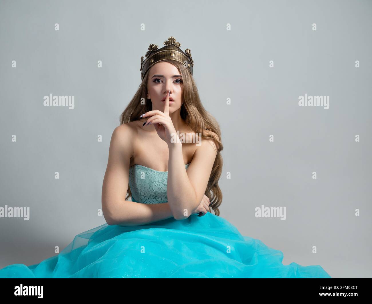 Fairy princess in a crown holds her finger to her lips, shhh, it's a secret. Young beautiful blonde in a lush blue dress. keep secrets, gray backgroun. Stock Photo