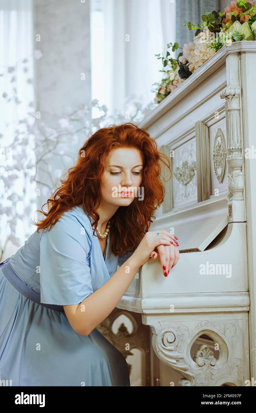 Beautiful red- haired young female pianist with blue eyes and freckles sits near a piano and learning music in studio. The creative process of making Stock Photo