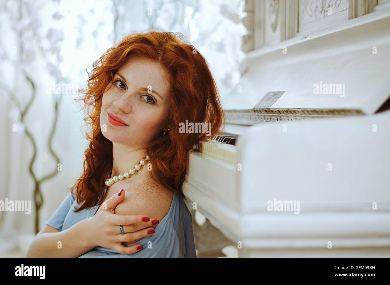 Beautiful red- haired young female pianist with blue eyes and freckles sits near a piano and learning music in studio. The creative process of making Stock Photo