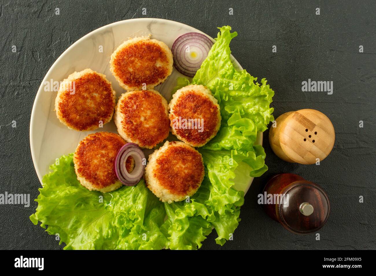 juicy delicious coated with breadcrumbs and fried chicken cutlets on white plate with spices in wooden shakers on dark background, view from above. Stock Photo