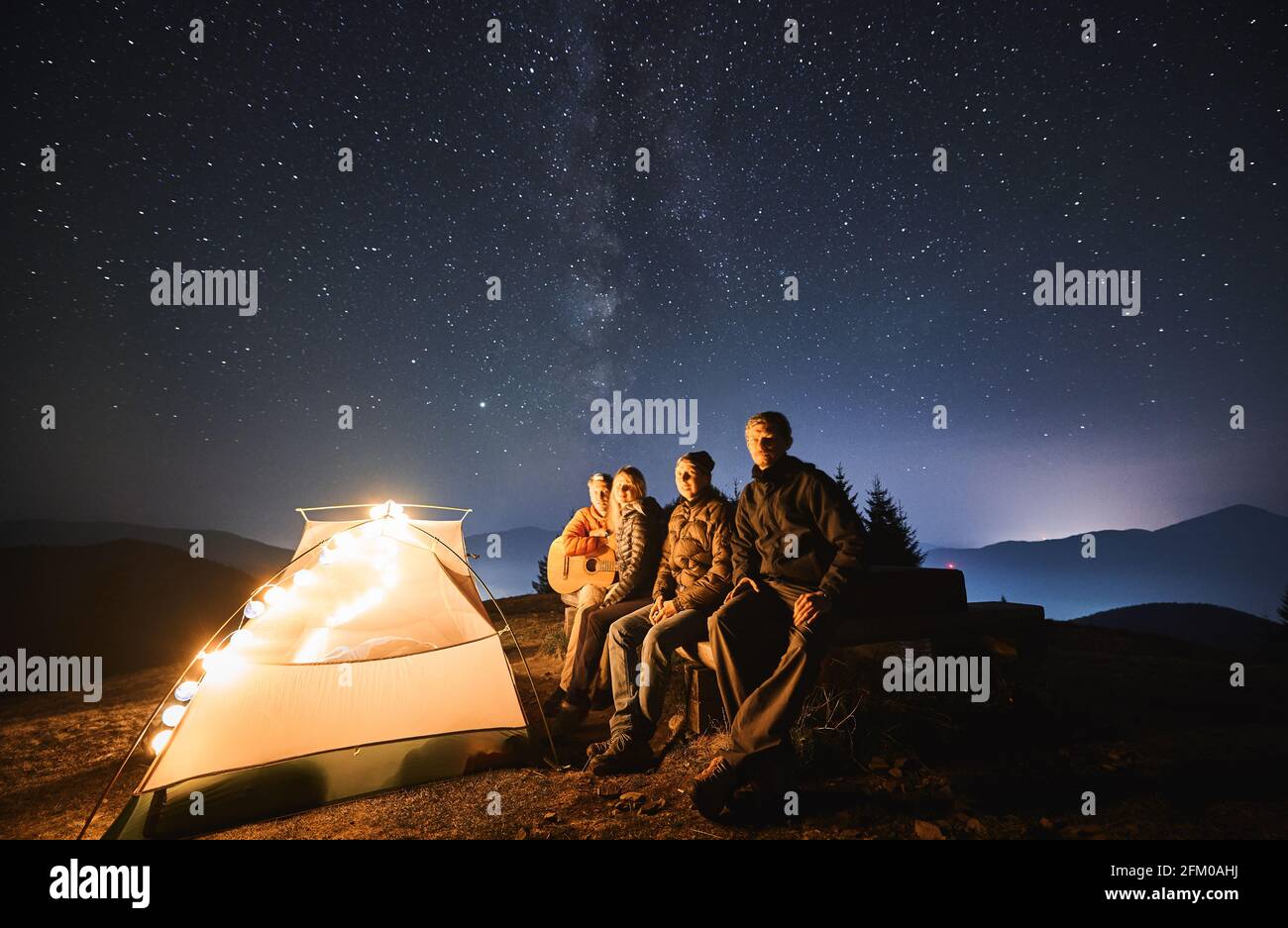 Hikers Sitting On Bench Near Illuminated Camp Tent Under Blue Night Sky With Stars Group Of Travelers Resting Near Tourist Tent Playing Guitar And Enjoying Beautiful View Of Night Starry Sky Stock