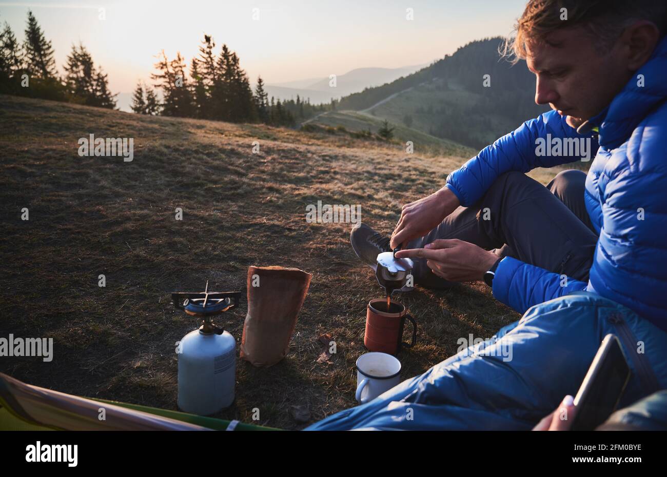 Handsome young man traveler sitting on grassy hill near camp tent and pouring hot drink into cup. Male hiker making coffee. Concept of hiking, camping and tourist equipment. Stock Photo