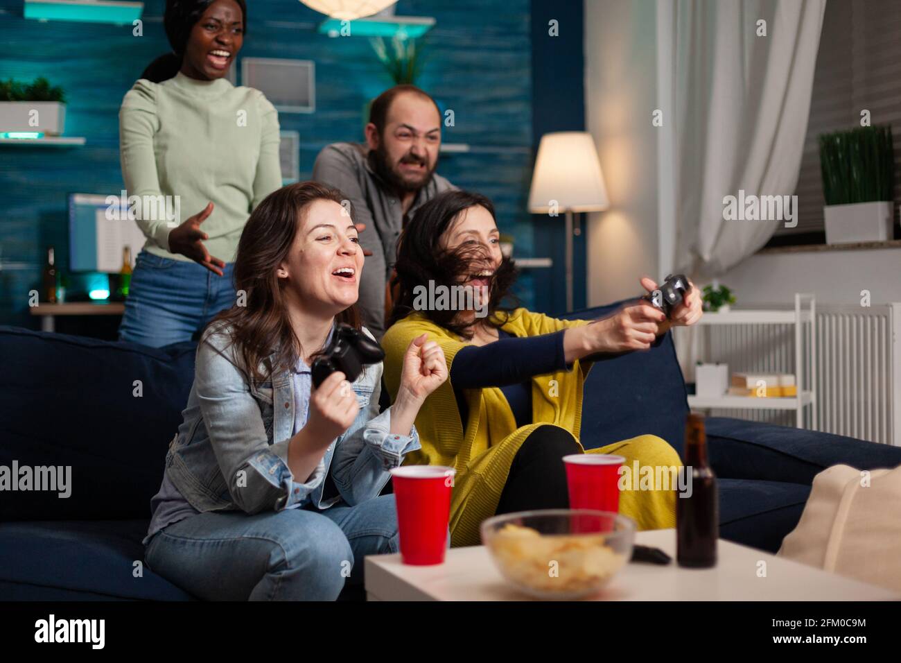 Multi ethnic group of people socializing winning at video games having fun. Group of mixed race friends playing games while sitting on sofa in living room late at night. Stock Photo