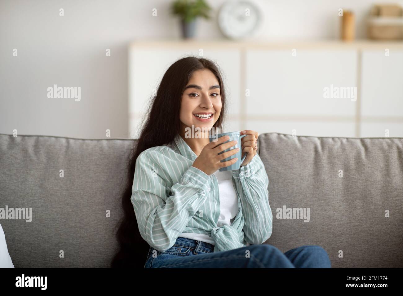 Portrait of pretty Indian woman chilling on couch with cup of aromatic coffee at home Stock Photo