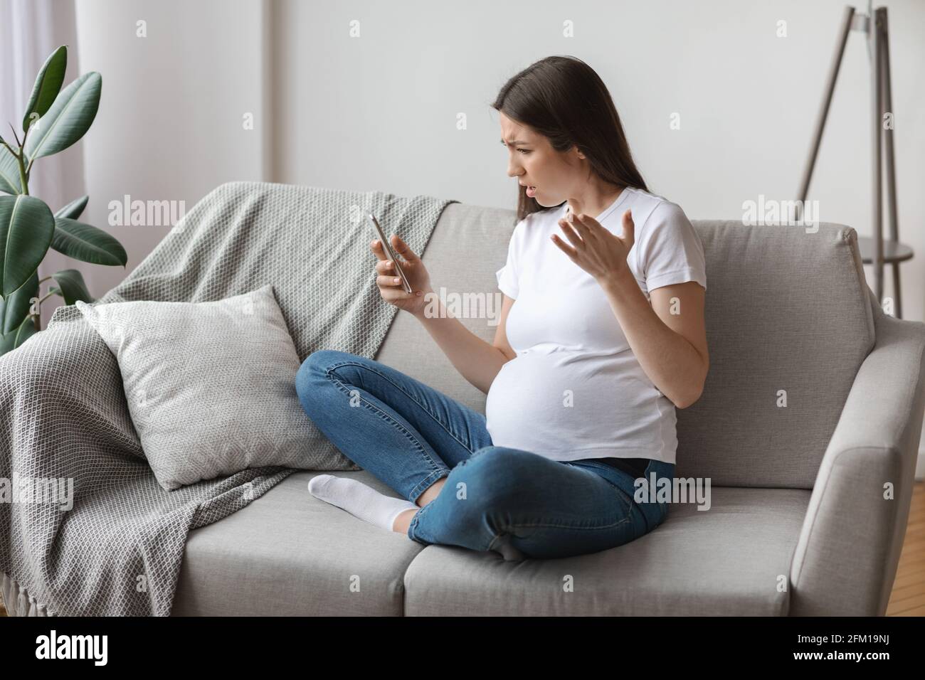Stressed Young Pregnant Woman Looking At Smartphone Screen While Relaxing On Couch Stock Photo