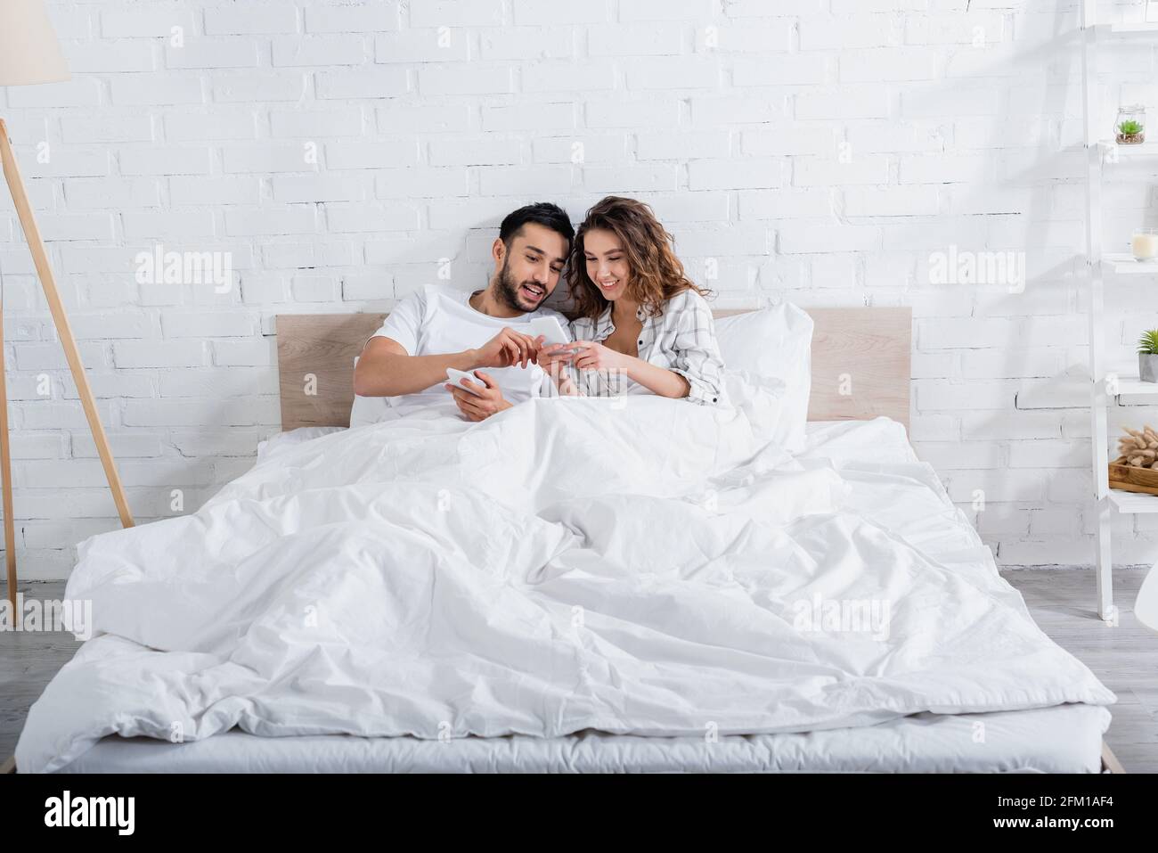 interracial couple smiling while lying on bed and using smartphones Stock Photo