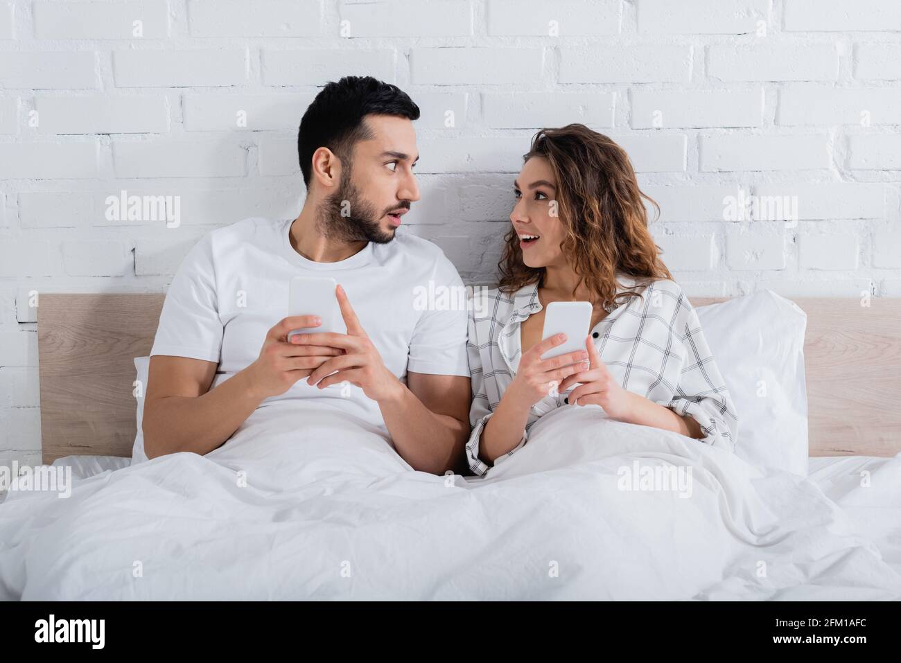 interracial couple looking at each other while lying on bed and using smartphones Stock Photo