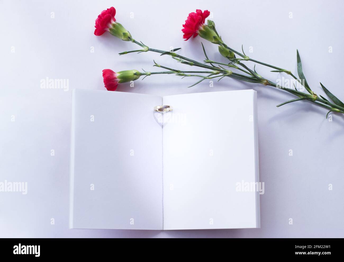 Summer wedding mockup. Blank wedding book on a white table. Wedding ring is placed on the book and the sunlight creates a heart-shaped shadow. Flowers Stock Photo