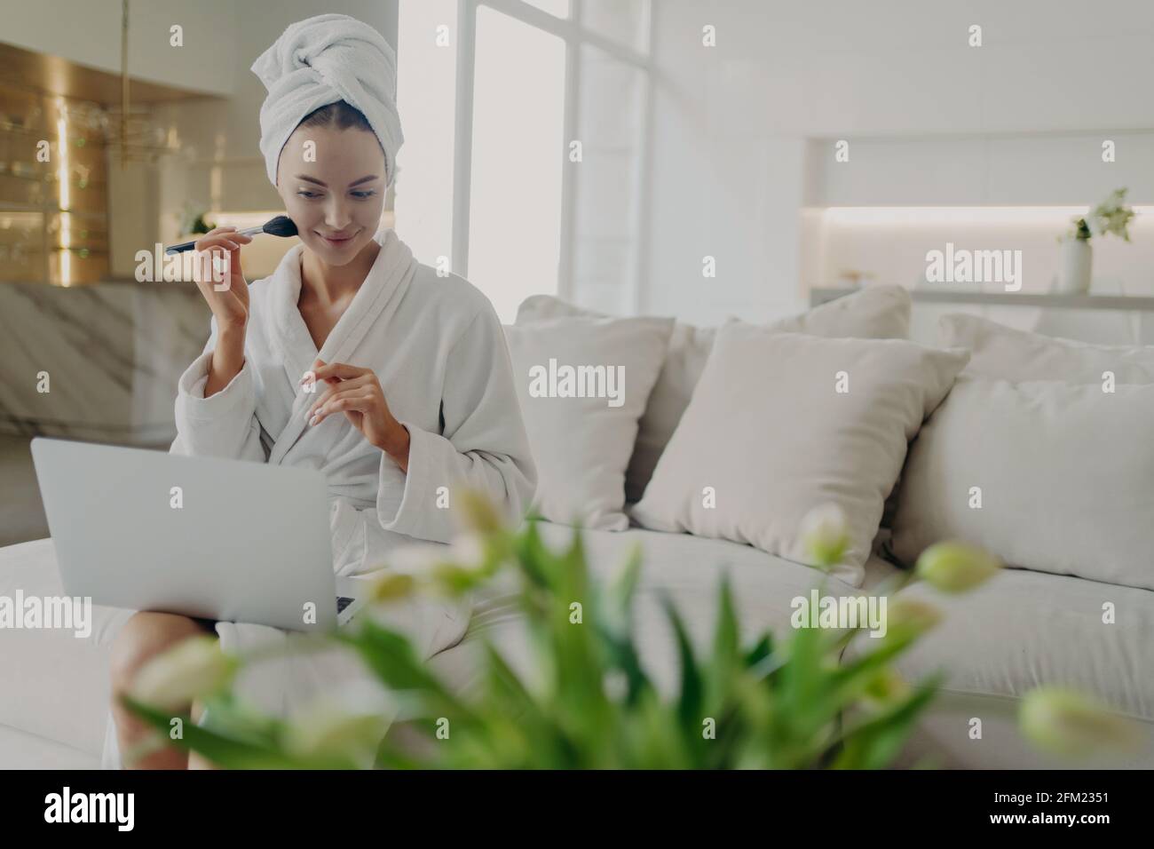 Young pretty woman in bathrobe doing daily makeup while watching video tutorial on laptop Stock Photo