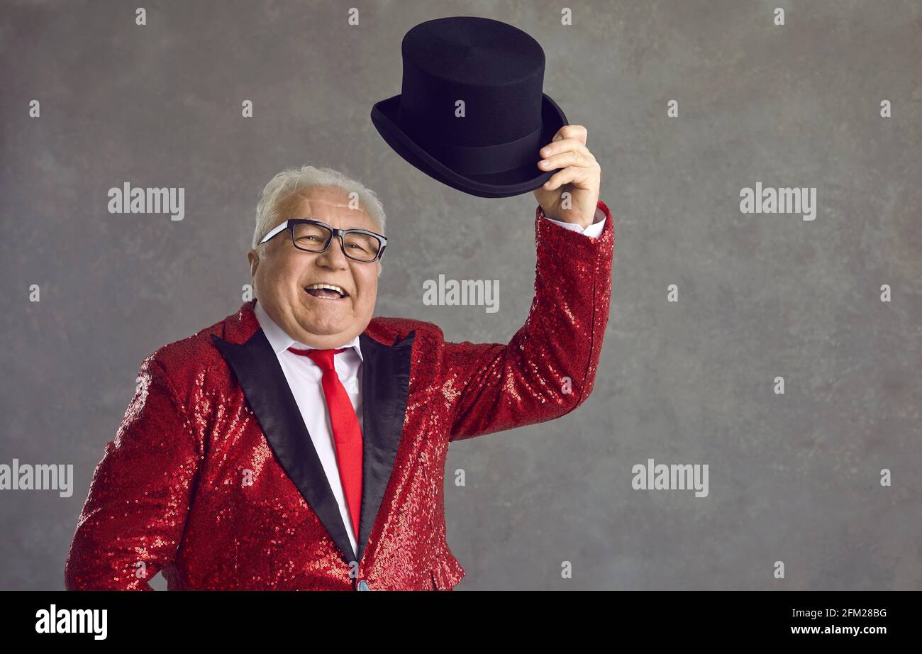 Happy senior presenter in red sequin jacket takes off his black top hat and greets audience Stock Photo