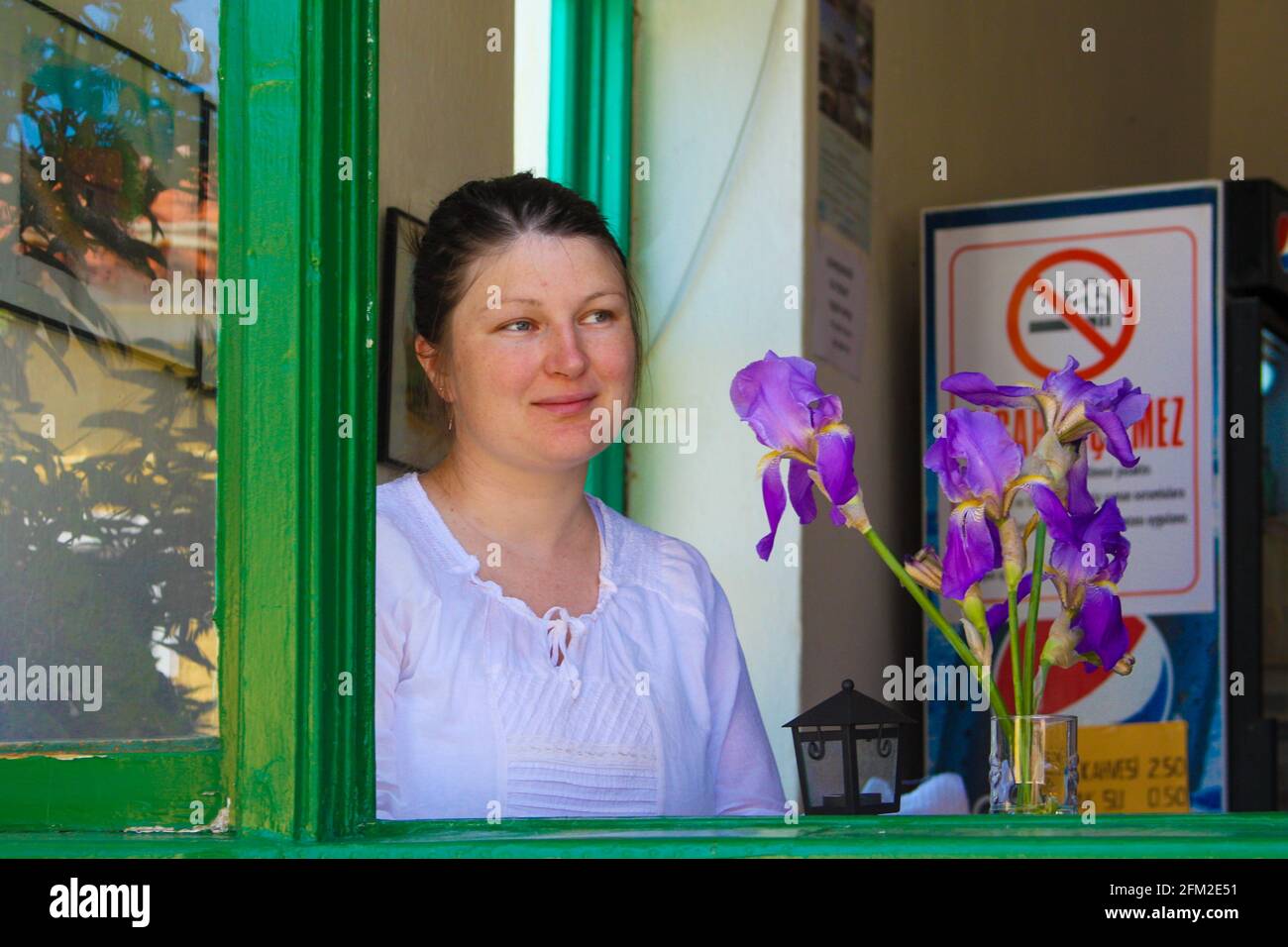 Gokceada, Canakkale - Turkey - May 06 2013: Young turkish woman wearing a white shirt, sitting at a cafe, looking out a green window, purple flowers i Stock Photo