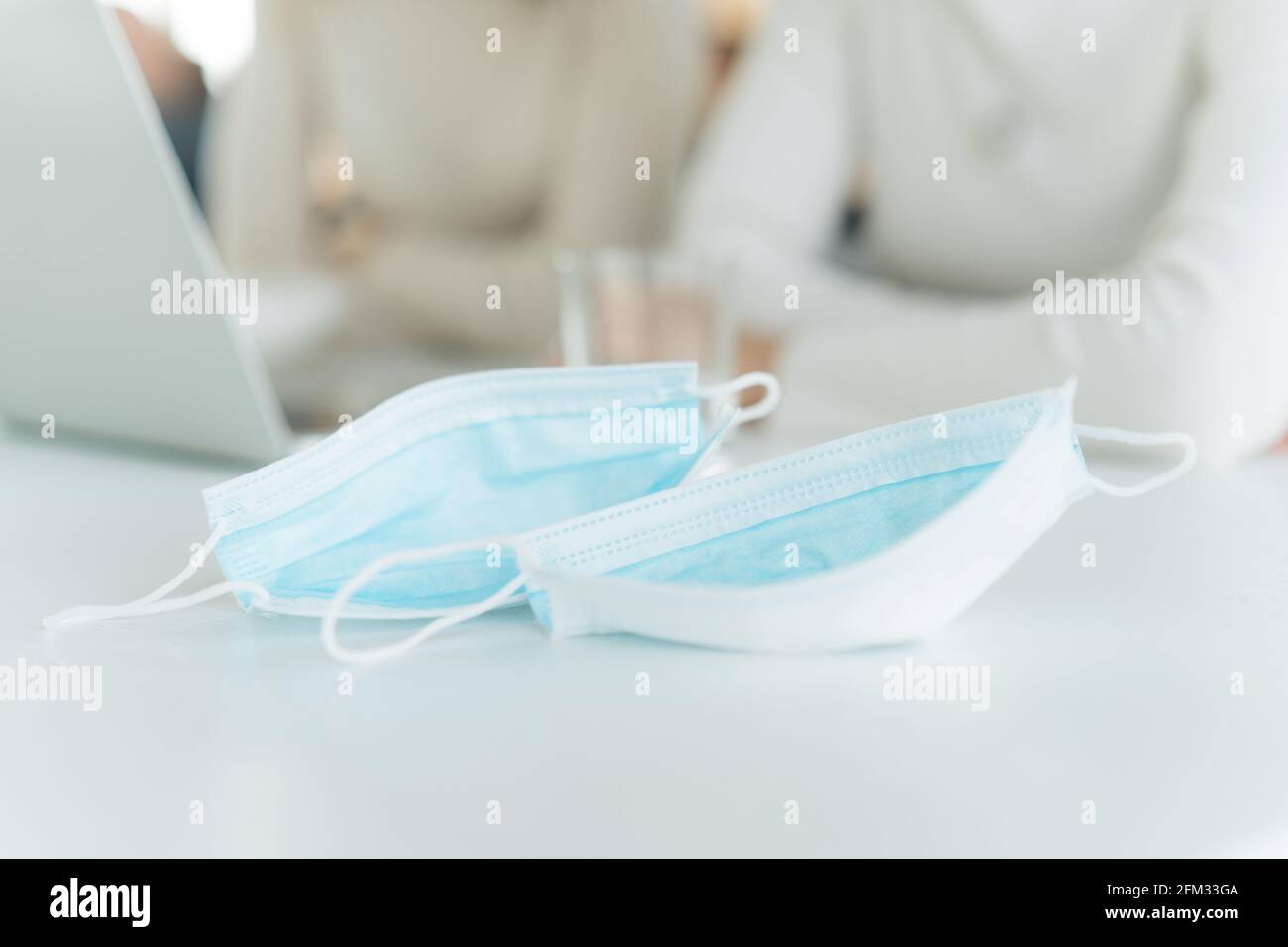 Close-up of two face masks on a table and two woman working on a laptop in background Stock Photo