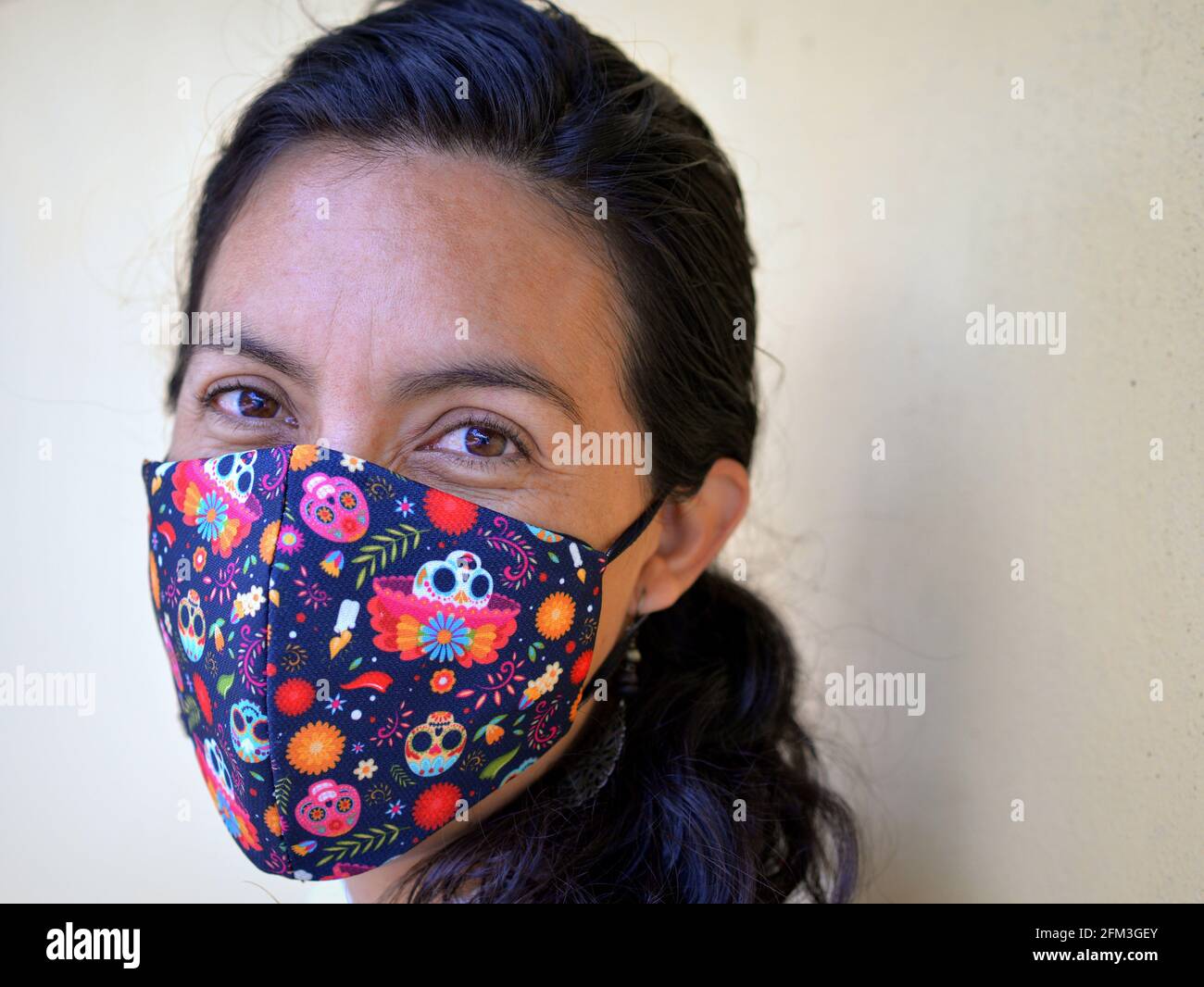 Young Mexican woman poses with stylish printed folkloric Dia-de-los-Muertos (Day of the Dead) cloth face mask during the global coronavirus pandemic. Stock Photo