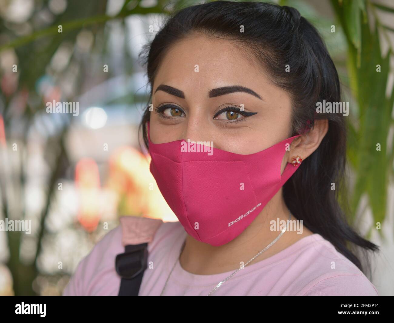 Beautiful young Latina woman with modern eye make-up and correct pink face mask poses for camera and keeps social distance during corona pandemic. Stock Photo