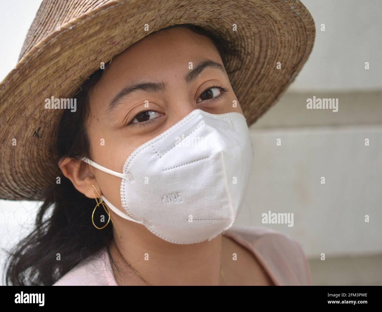 Brown-eyed young Mexican woman wears a worn Panama summer straw hat and a white KN 95 protective face mask during the global coronavirus pandemic. Stock Photo