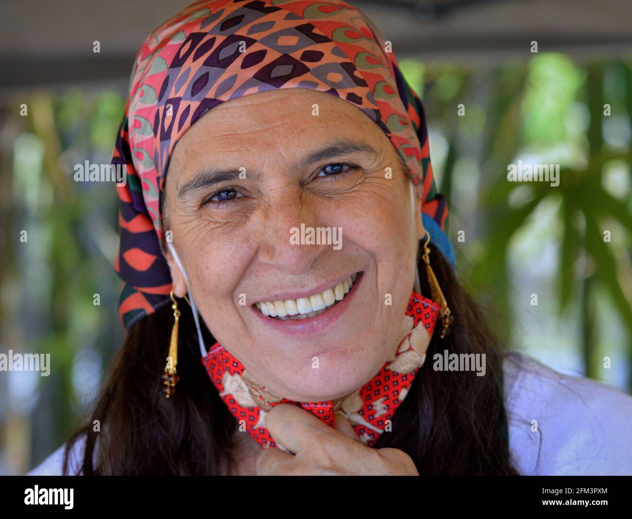 Mature positive Caucasian countrywoman (gardener) wears a modern head scarf and pulls down her red cloth face mask for a natural genuine teeth smile. Stock Photo