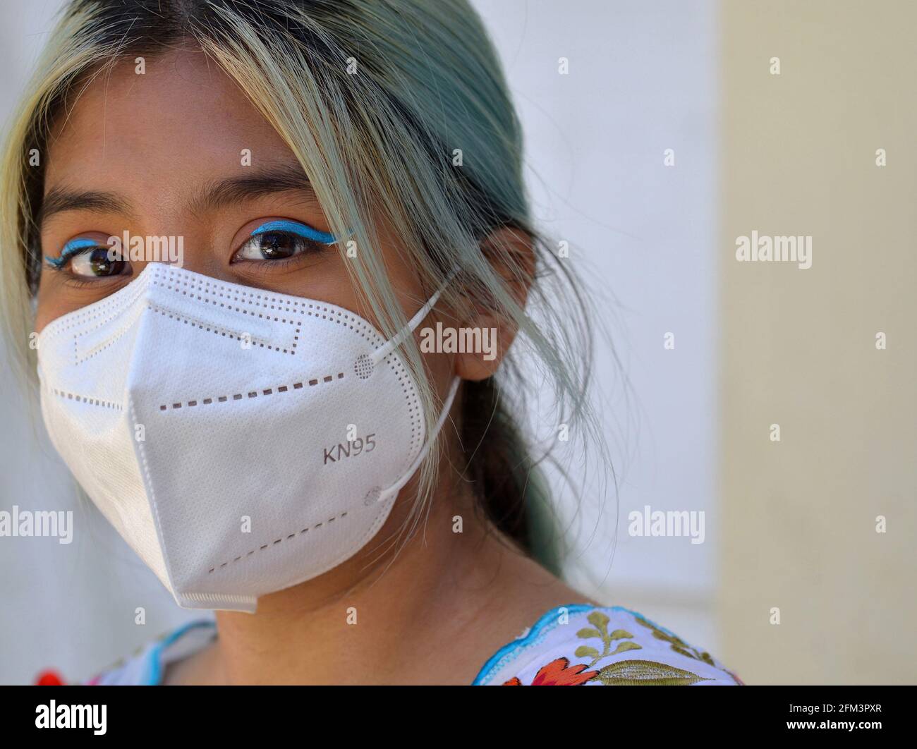 Mexican teen girl with dyed hair, big brown eyes and elaborate blue eye makeup wears a white KN95 face mask during the global coronavirus pandemic. Stock Photo