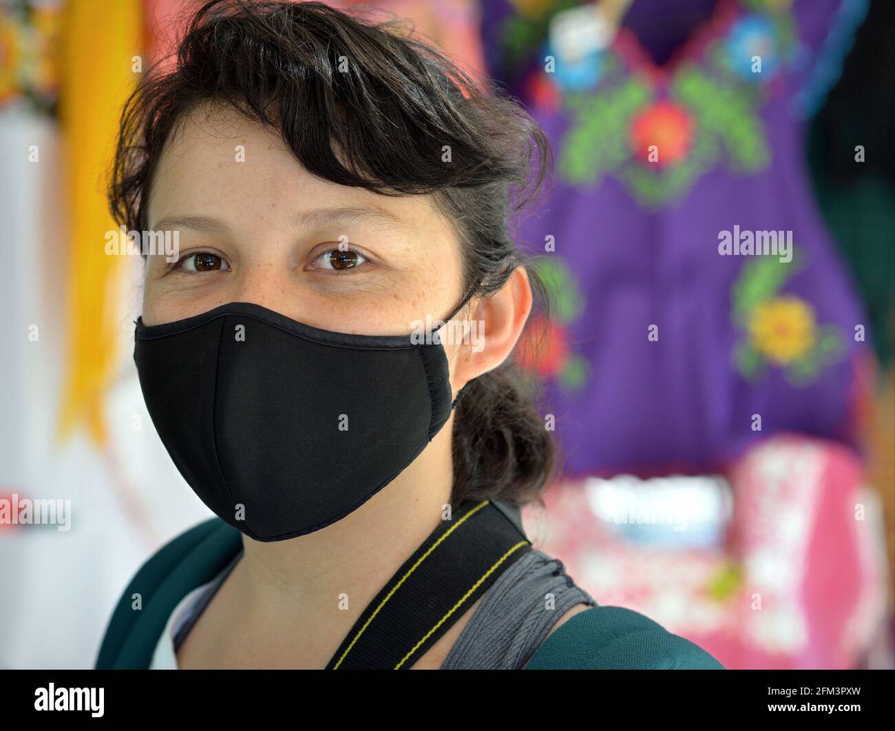 Young Mexican woman wears a black face mask during the global coronavirus pandemic and poses for the camera in front of a folkloric clothes stall. Stock Photo