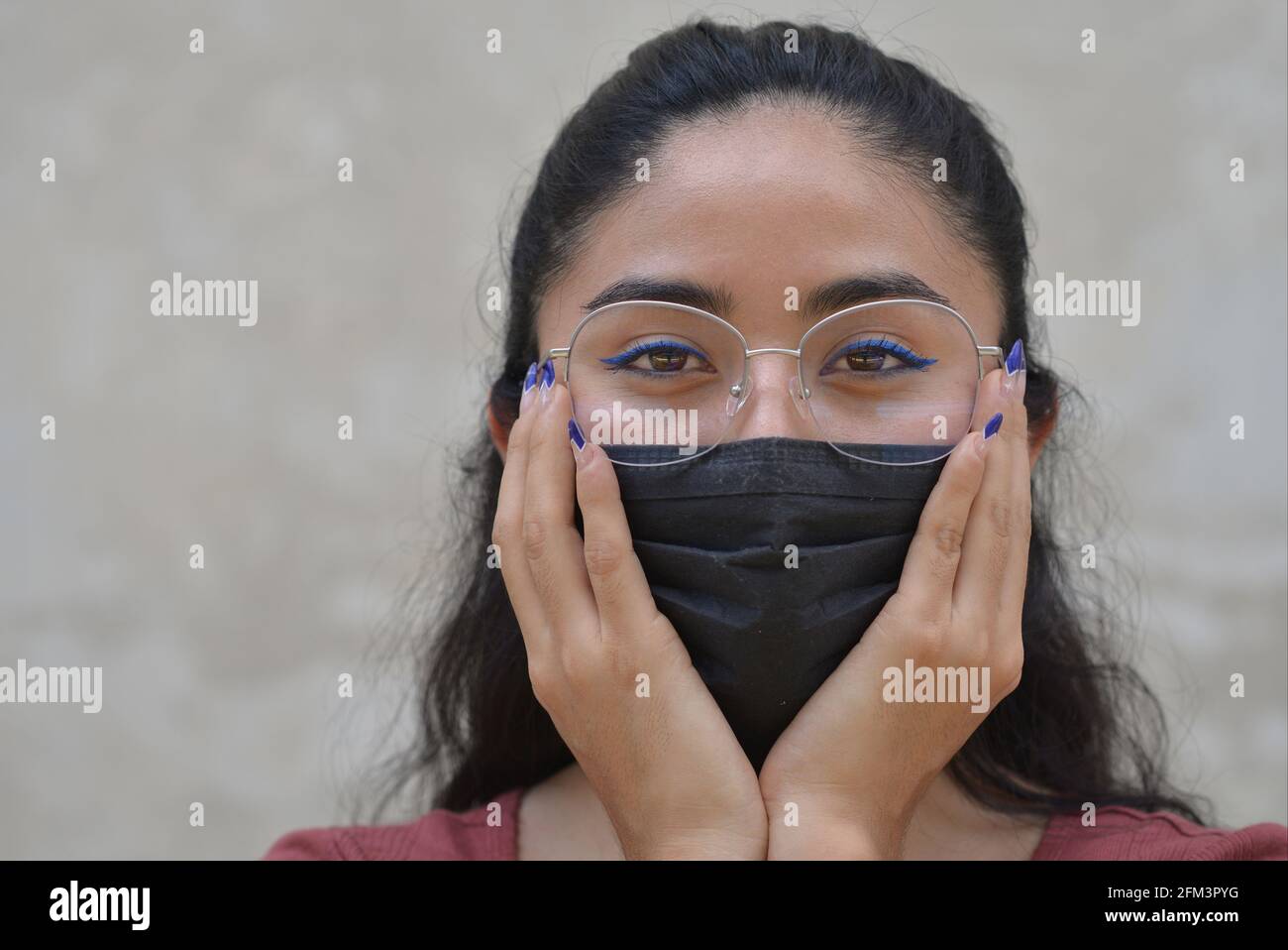 Young stylish beautiful Mexican woman with blue eyeliner and blue nail enamel wears eyeglasses and a black pandemic face mask and looks at the viewer. Stock Photo