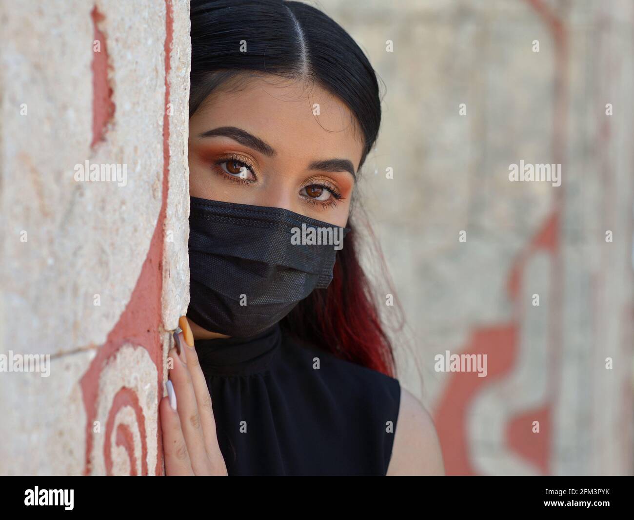 Beautiful Caucasian Mexican model with eye makeup and black outfit wears a black medical face mask and looks around the corner of a stone wall. Stock Photo