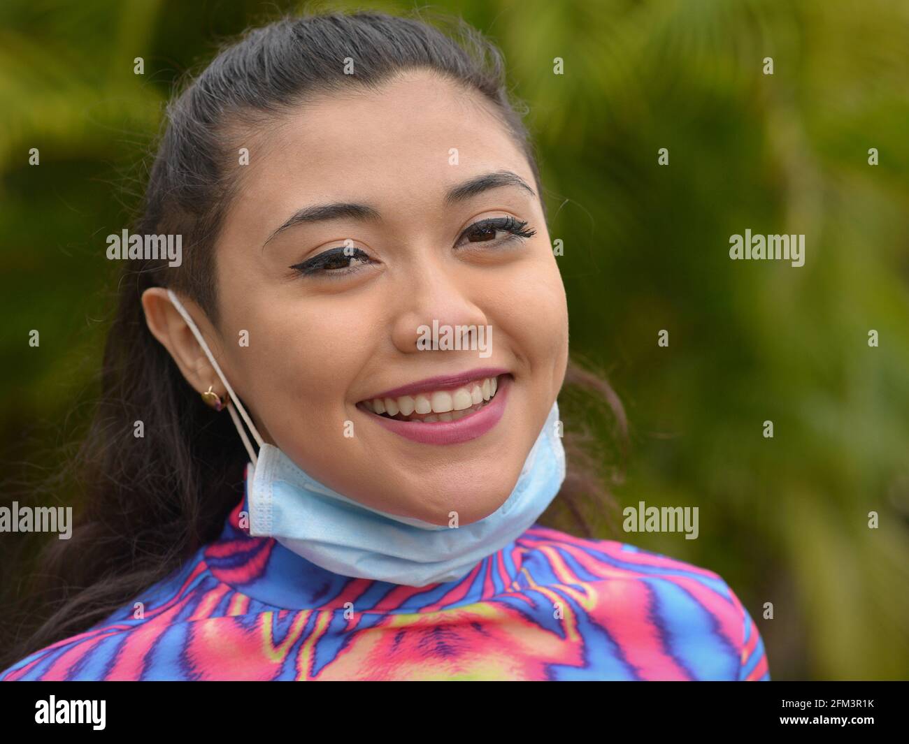 Young optimistic pretty Caucasian woman with eye makeup pulls down her light-blue surgical ear loop face mask and smiles for the camera. Stock Photo