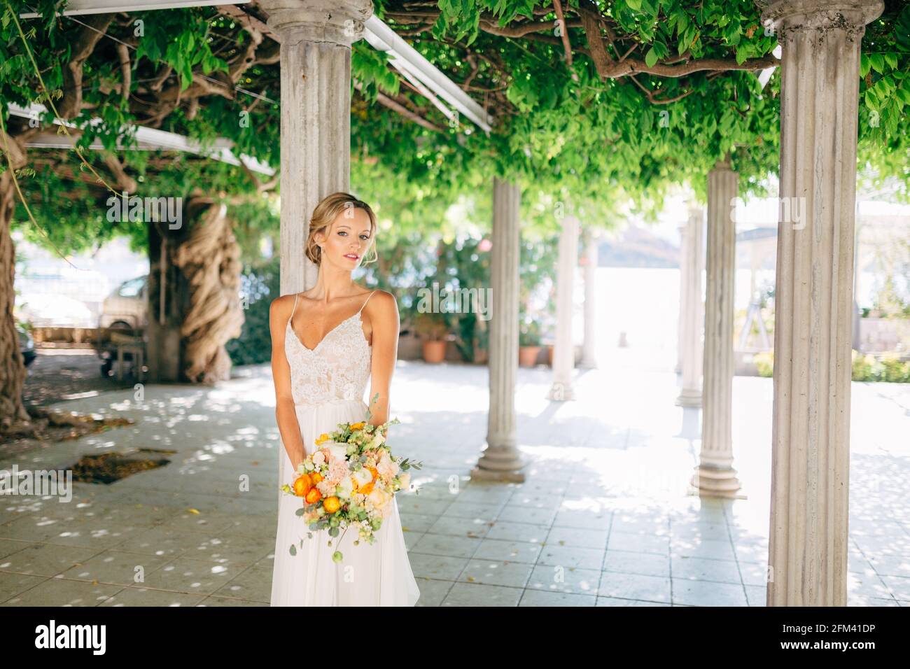 The bride stands among white columns braided with green vines and holds a  wedding bouquet with orange buttercups, close-up Stock Photo - Alamy