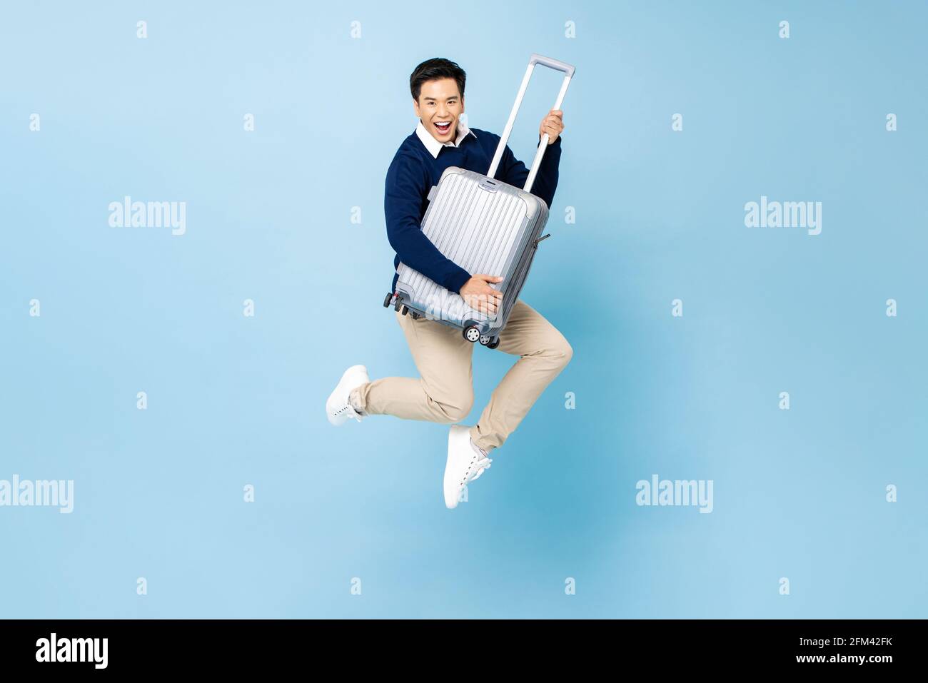 Fit healthy smiling young handsome Asian tourist man with baggage jumping in mid-air ready to fly isolated on light blue studio background Stock Photo