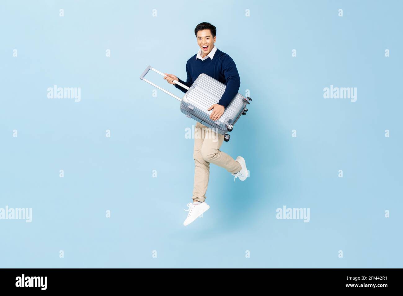 Jumping portrait of young fit healthy smiling handsome Asian tourist man with baggage ready to fly isolated on light blue studio background Stock Photo