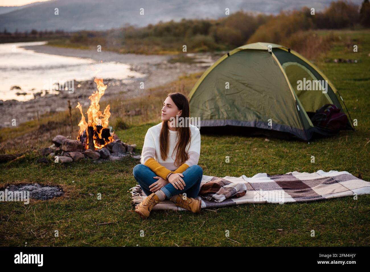 Front view of young brunette in the afternoon sits near a tent and enjoys mountain views. The campfire is lit on the side. Camping and hiking in the m Stock Photo