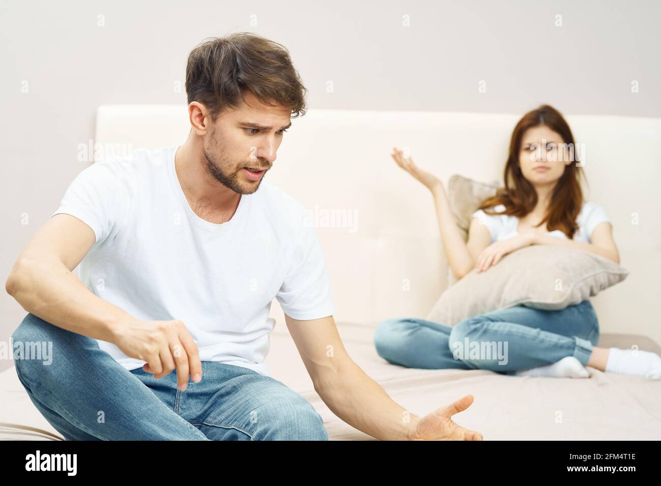 a man in a white t-shirt and jeans communicates with a woman on the bed in the background Stock Photo