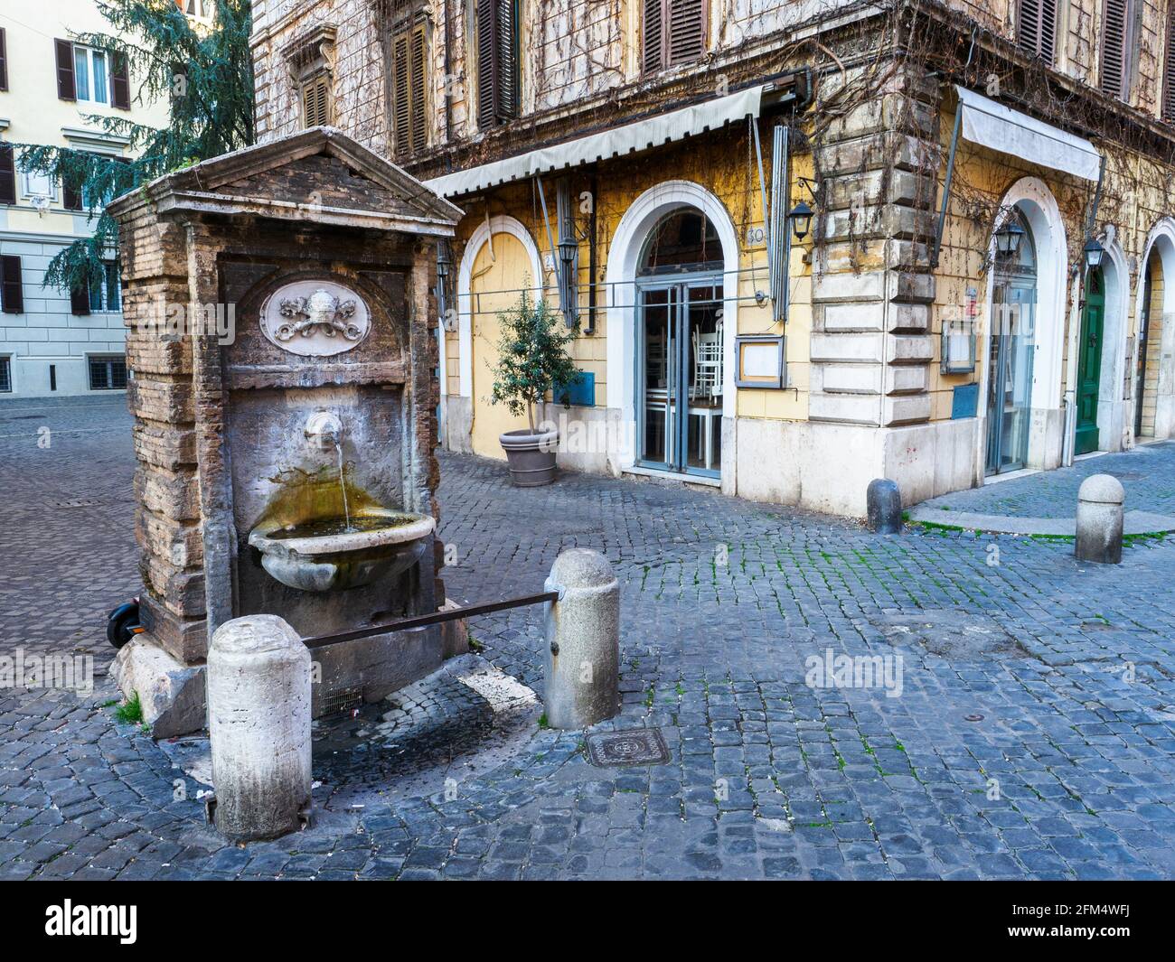 The fountain  in rione Borgo is protected by two small columns connected by an iron bar, consists of a simple rectangular travertine aedicule, gable at the top and leaning against a brick construction. The upper arch has a papal coat of arms - Rome, Italy Stock Photo