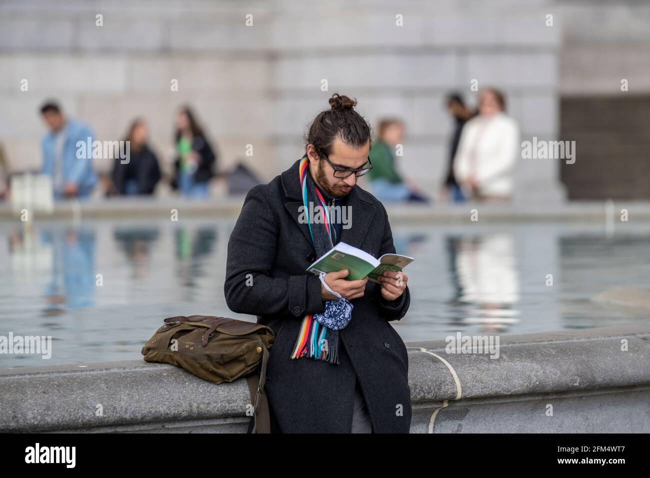 Candid in public park. A generation z man is seen reading a book on the edge of a water fountain. With black hair tied back in a bun and glasses. Stock Photo