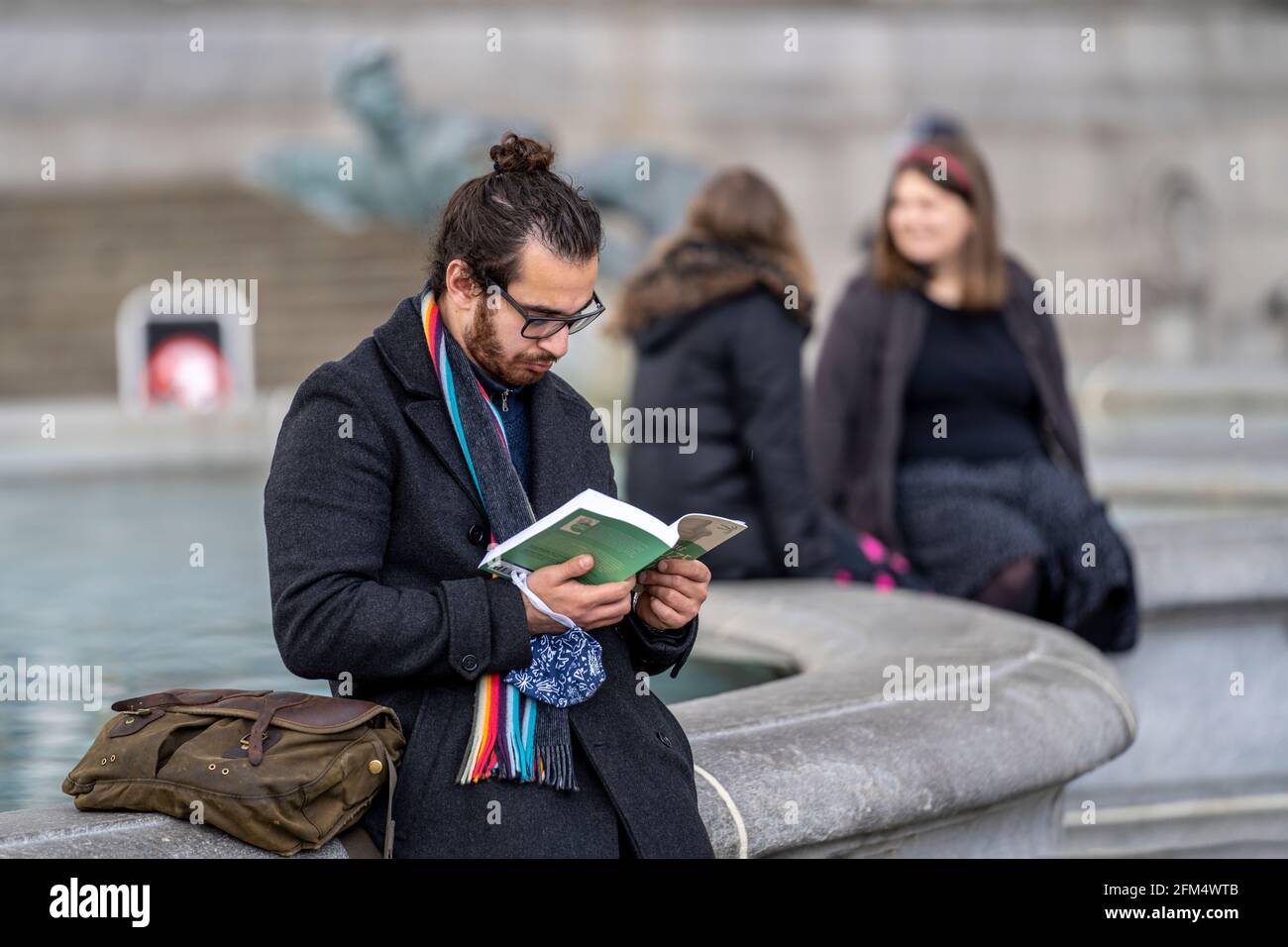 Candid in public park. A generation z man is seen reading a book on the edge of a water fountain. With black hair tied back in a bun and glasses. Stock Photo