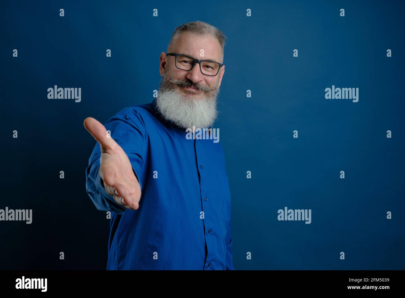 Mature bearded caucasian man holds out his hand to say hello. Friendly grayhaired bearded senior man dressed blue shirt makes greeting gesture over bl Stock Photo