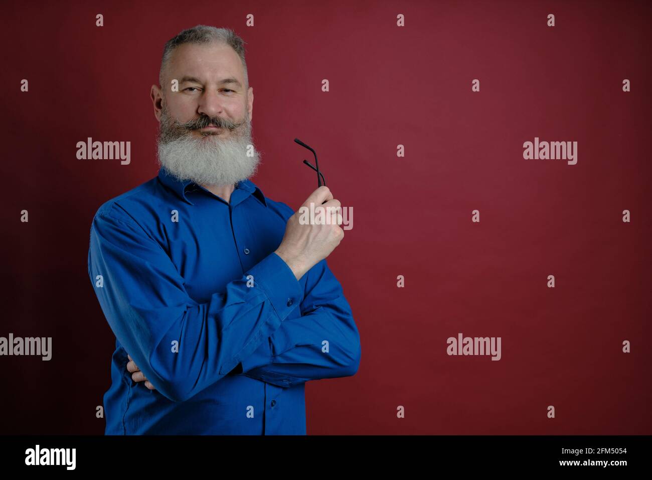 Mature bearded caucasian man dressed blue shirt looks confident at camera with eyeglasses in hand, red backdrop Stock Photo