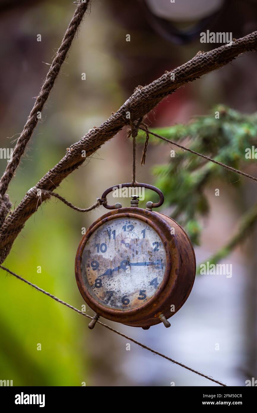 Old and rusty alarm clock hung in the garden as decoration. Photo taken in low light conditions. Blurred background. Stock Photo