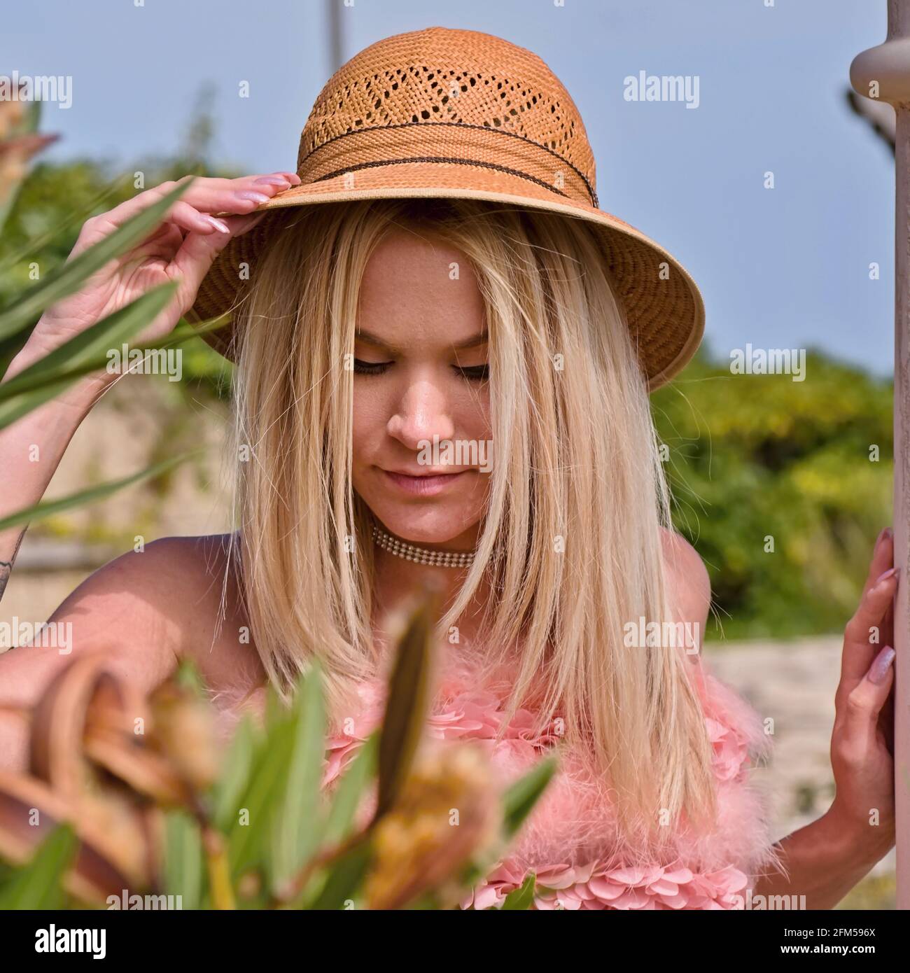 A young girl with long blonde hair is stnding in a flower bed. She is wearing a sun hat and her right arm is over the hat, looking down. She waers a r Stock Photo