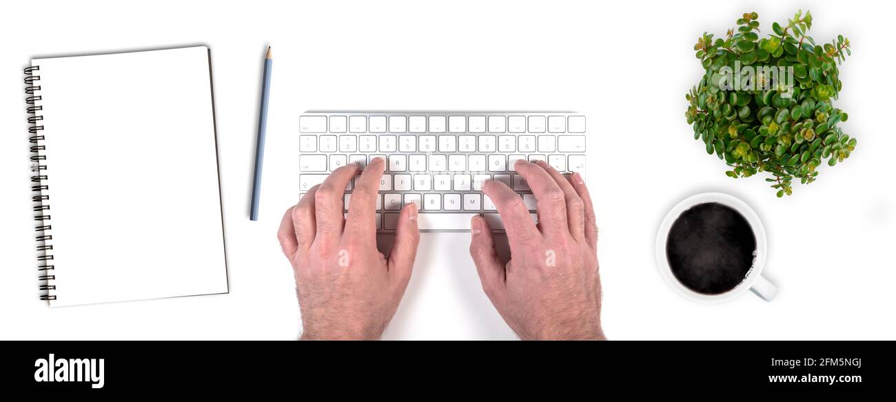 top view of person typing on wireless computer keyboard on white desk with potted plant, notepad with pencil and cup of coffee Stock Photo