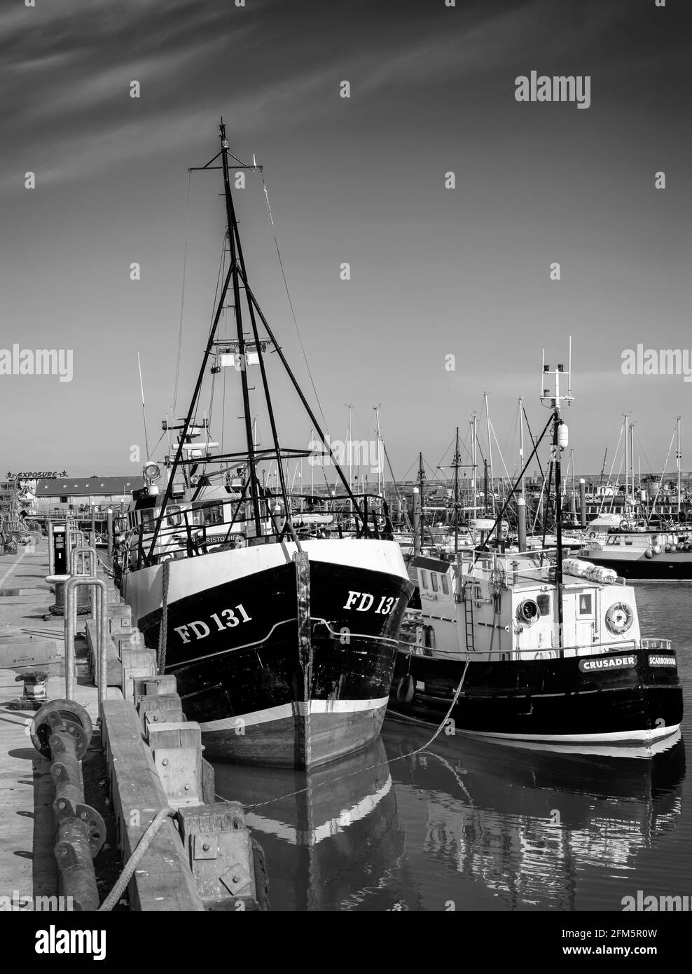 Trawlers are moored alongside a wharf with reflections in the water.  The yachts of a marina are beyond and a clear sky is above. Stock Photo