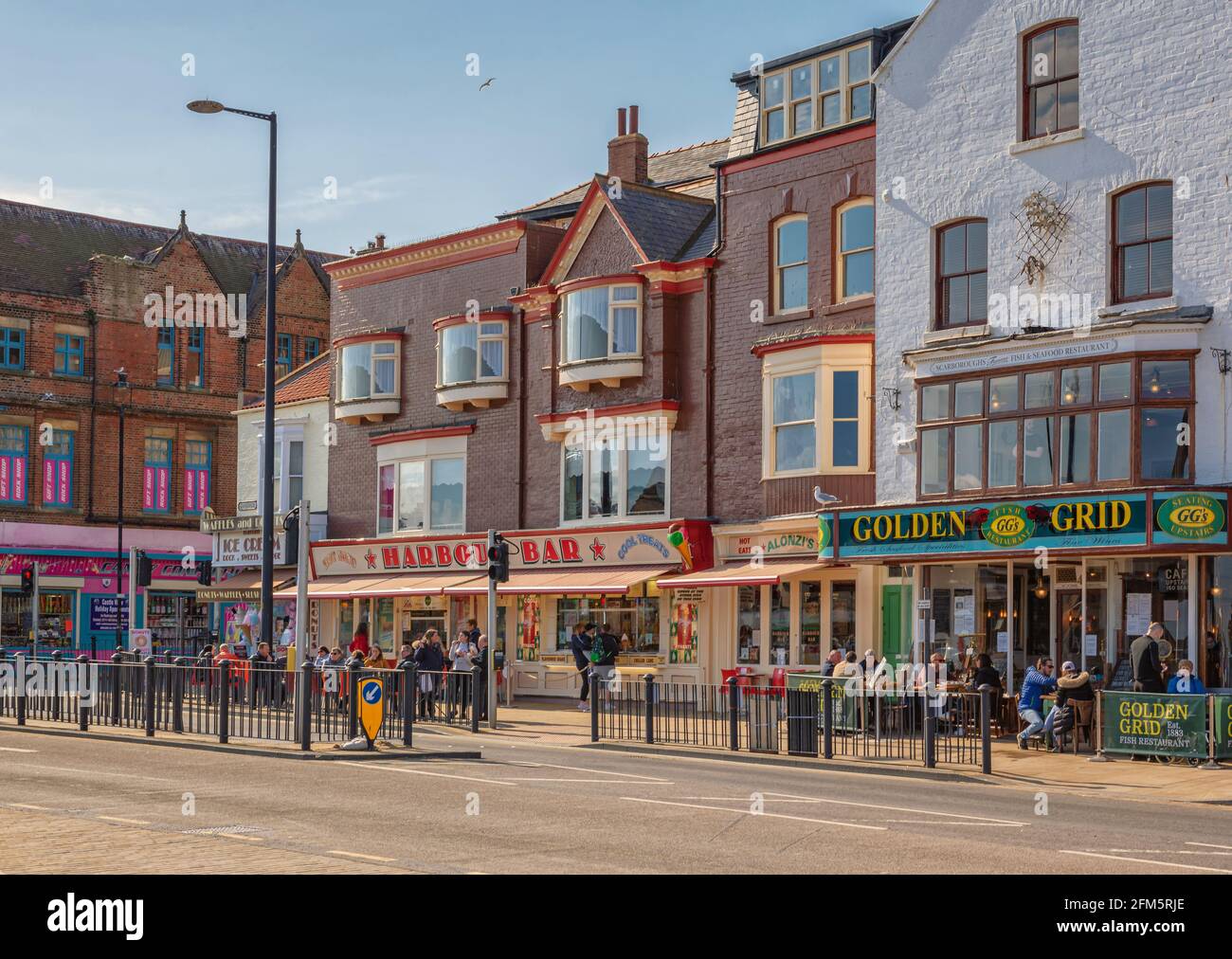 Seafront road with a restaurant, bar and ice cream kiosk.  People sit and stand in the sunshine and a clear sky is overhead. Stock Photo